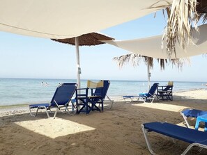 A beach with lounge chairs under a canopy, a clear sky, and the sea in the background. - Villa- Nea Kalikrateia - 2-bedroom Apartmen (Nea Propontida)