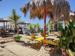 Ein Strand mit Liegestühlen, Reetdachsonnenhütten und Blumentöpfen.