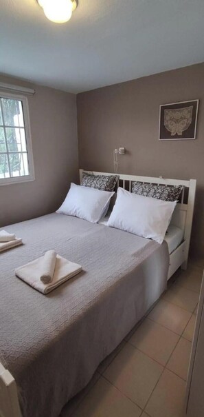 A neatly made bed with a white headboard, a window with blinds, and a wall-mounted light fixture. - Villa- Nea Kalikrateia - 2-bedroom Apartmen (Nea Propontida)