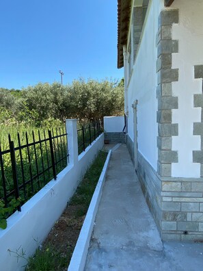 A narrow concrete pathway leading to a building with a stone and white facade, bordered by a black metal fence and greenery. - Villa- Nea Kalikrateia - 2-bedroom Apartmen (Nea Propontida)