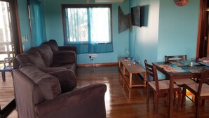 Una sala de estar con un sofá, una mesa de comedor con sillas, un televisor montado en la pared y una ventana con cortinas.