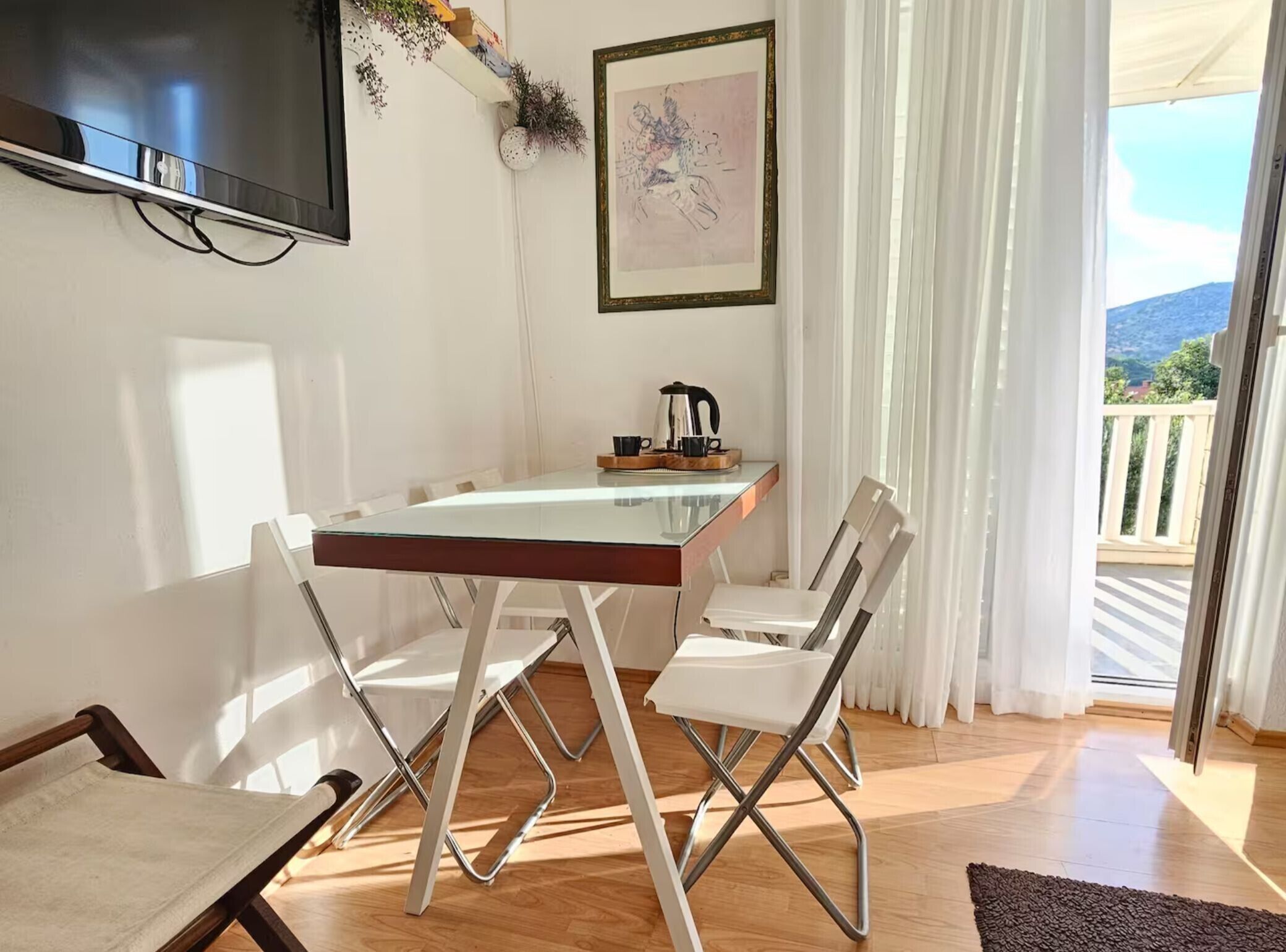 A room with a glass-top table, white chairs, a flat-screen TV mounted on the wall, a framed artwork, and a view of a balcony with a railing.