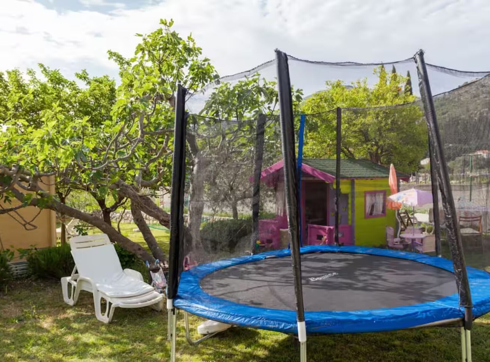 Ein Trampolin mit Sicherheitsnetz, ein weißer Liegestuhl und eine kleine, pink- und grün gefärbte Konstruktion in einem Garten.