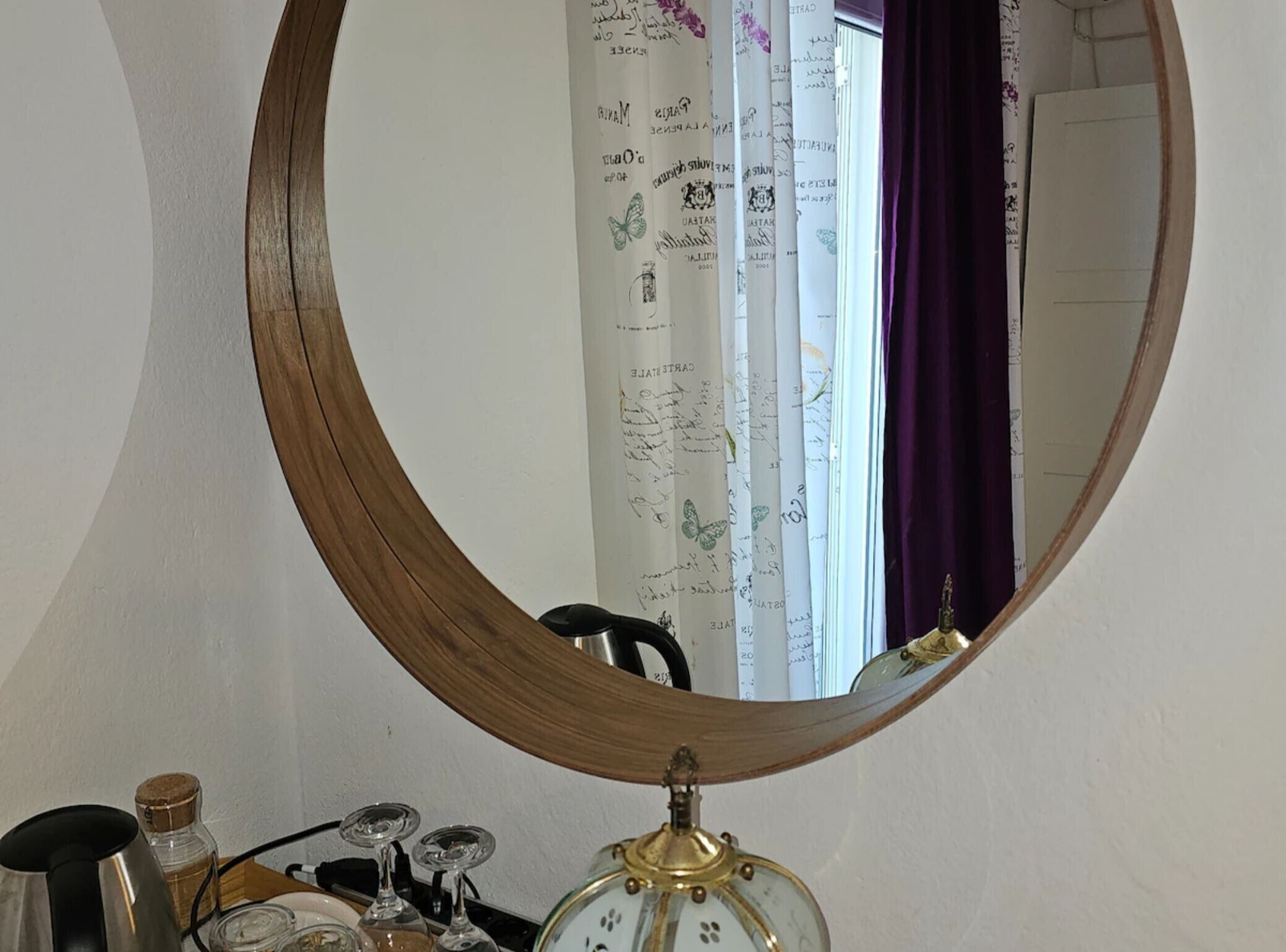 A round mirror reflecting a room with a window, curtains, a clock, and a small table with items.