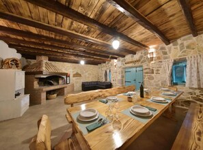 Un coin repas rustique avec une longue table en bois dressée pour le repas, des murs en pierre et une cheminée en pierre.