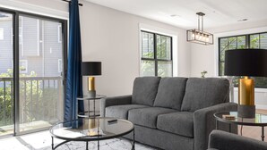 A modern living room with a grey sofa, a glass coffee table, and a large window with a view of greenery. - Luxury Modern Condo Near Notre Dame 2BD| 2BA (South Bend)