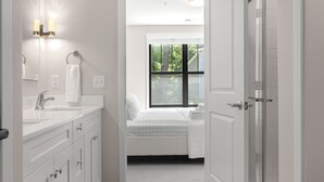 A modern bathroom with a white vanity, a mirror with wall sconces, a shower area, and a window with a view of trees. - Luxury Modern Condo Near Notre Dame 2BD| 2BA (South Bend)