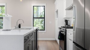 A modern kitchen with a central island, stainless steel appliances, and a window with a view of greenery. - Luxury Modern Condo Near Notre Dame 2BD| 2BA (South Bend)