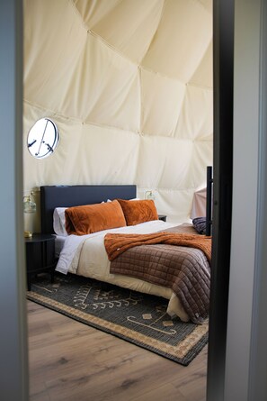 Un dormitorio con cama, almohadas, mesita de noche, lámpara y alfombra.