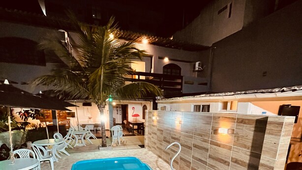 Piscina com deck ladrilhado, rodeada por cadeiras e mesas brancas, e uma palmeira.