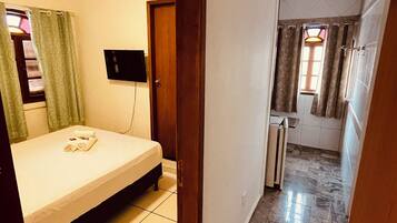 A hotel room with a bed, a TV, a bathroom with a shower, and a hallway leading to another room.