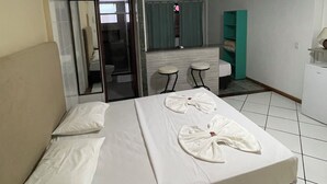 Quarto de hotel com cama, escrivaninha, cadeira, ventoinha no teto e banheiro visível através de uma porta de vidro.