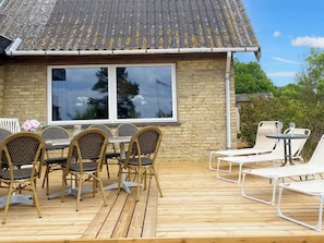 Eine Holzterrasse mit Korbstühlen und einem Tisch, ein Backsteinhaus mit einem großen Fenster und ein weißer Liegestuhl.