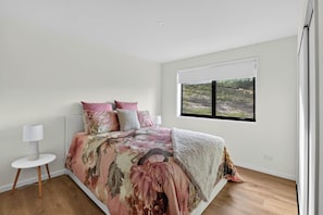 A bedroom with a large bed, a window with a view of a natural landscape, a bedside table with a lamp, and a white wall.