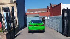 Ein grünes Auto parkt auf einer Auffahrt mit einer blauen Garagentür, einer Ziegelwand und einem Metalltor.