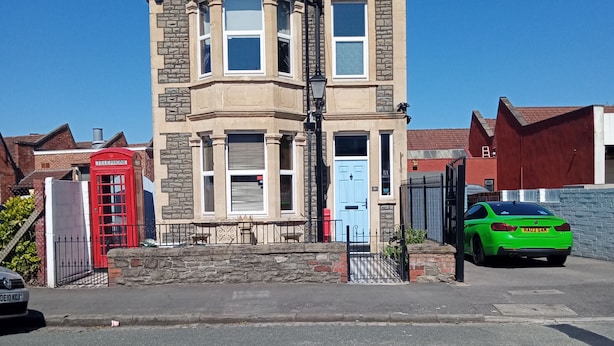 Ein zweistöckiges Haus mit einer Stein-Fassade, eine rote Telefonzelle und ein grünes Auto parken davor.