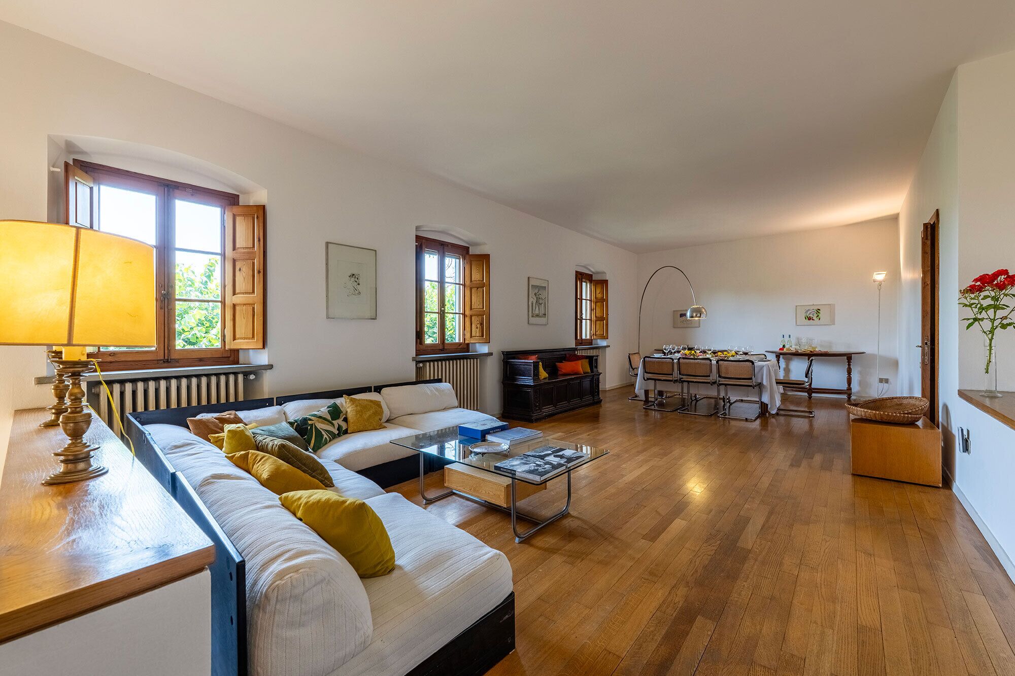 A spacious living area with a sofa, dining table, and chairs, wooden floors, and large windows.