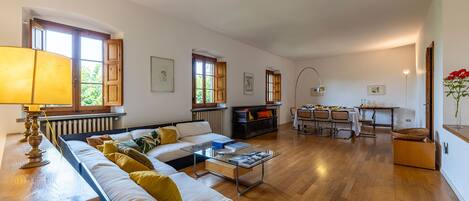 Amplia sala de estar con sofá, mesa de comedor y sillas, pisos de madera y ventanas grandes.