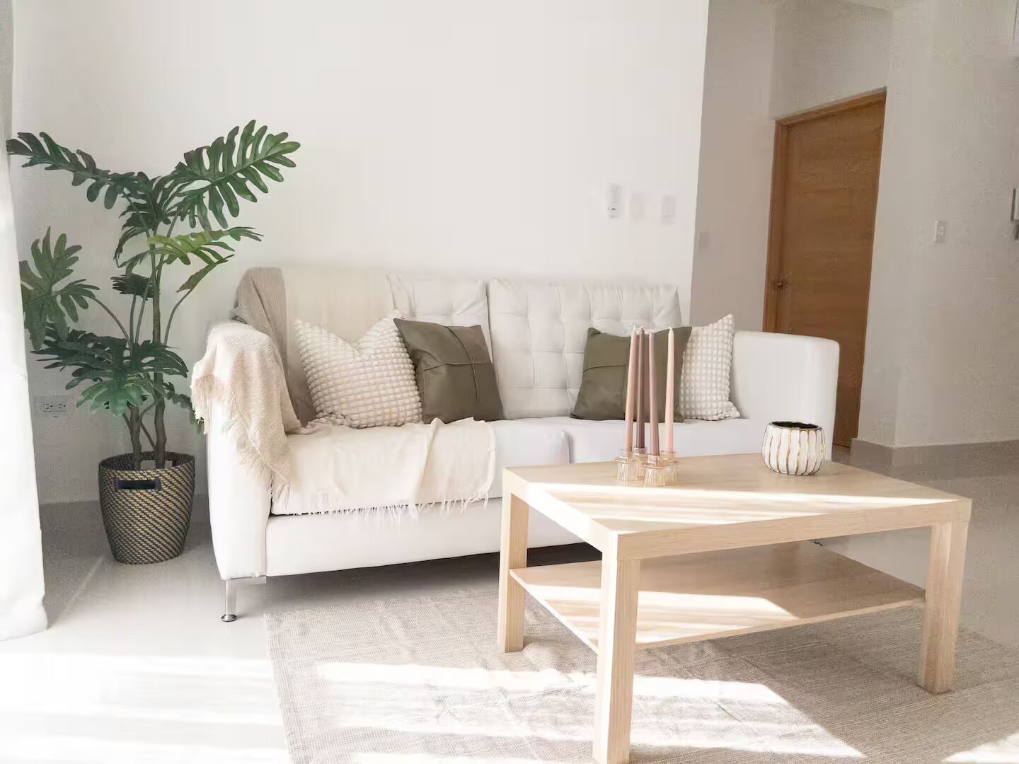 Un canapé blanc agrémenté d’un plaid beige, une plante en pot et une table basse en bois, le tout dans une pièce lumineuse où repose un tapis à motifs.