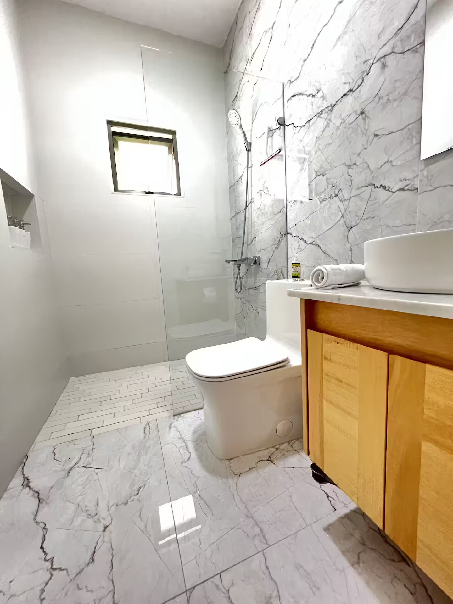 A bathroom with a toilet, shower, marble floor, and wooden vanity.