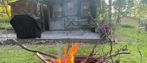 A rustic outdoor setting with a fire pit, a covered grill, and a house with a porch.