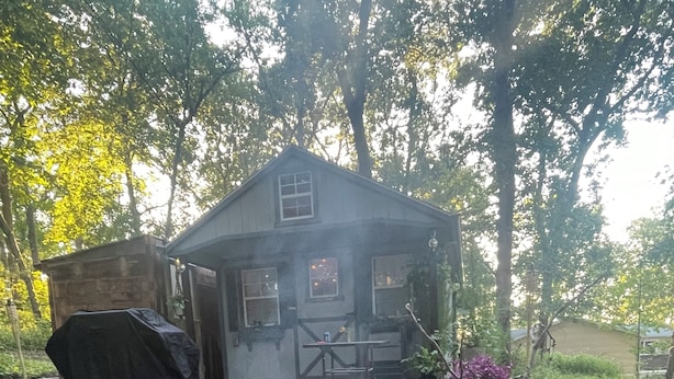 Un cadre extérieur rustique avec un foyer, un barbecue couvert et une maison dotée d’un porche.