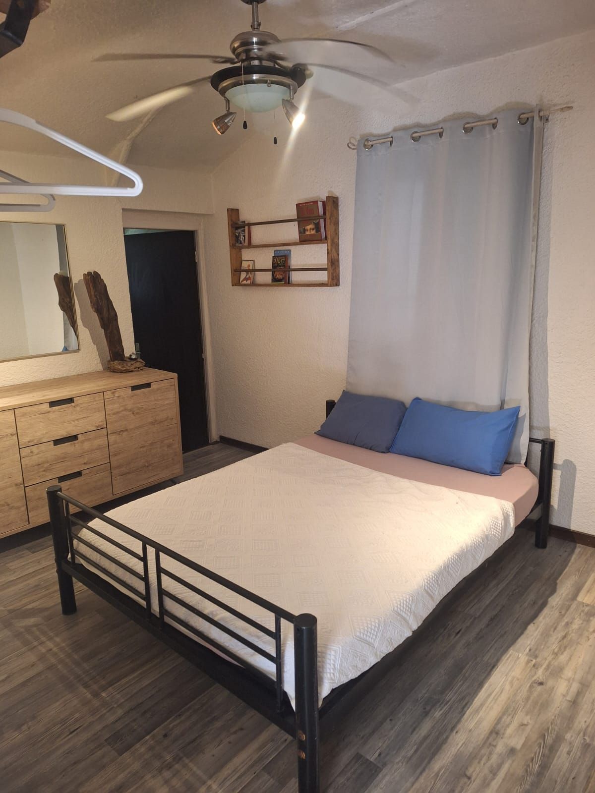 A bedroom with a bed, a dresser, a ceiling fan, and a wall-mounted shelf with books.