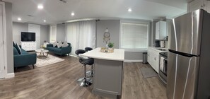 A modern kitchen with a stainless steel refrigerator, a dining area with a white table and bar stools, and a living room with a teal sofa and a flat-screen TV. - Luxury suite in Bellingham, MA (Bellingham)