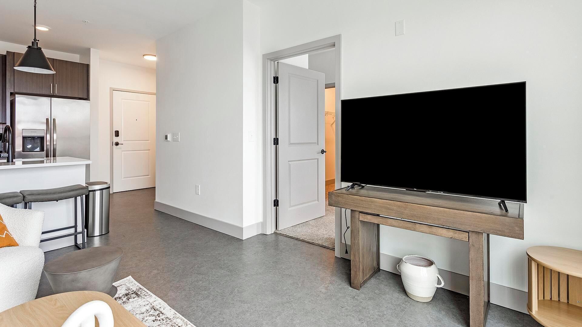 A modern living room with a large flat-screen TV, a kitchen with a built-in fridge, and a dining area with bar stools.