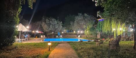 A nighttime outdoor pool area with a tiled pathway, lounge chairs, and ambient lighting.