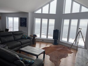 A living room with a leather sofa, a coffee table, a large window with a view of a body of water, and a tripod with a camera.