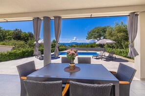 Ein überdachter Essbereich im Freien mit Blick auf einen Pool, Korbstühle und ein Tisch mit einer Vase.
