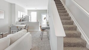 Een moderne keuken met witte kasten, een eethoek met een grijze tafel en stoelen, en een wenteltrap met tapijt.