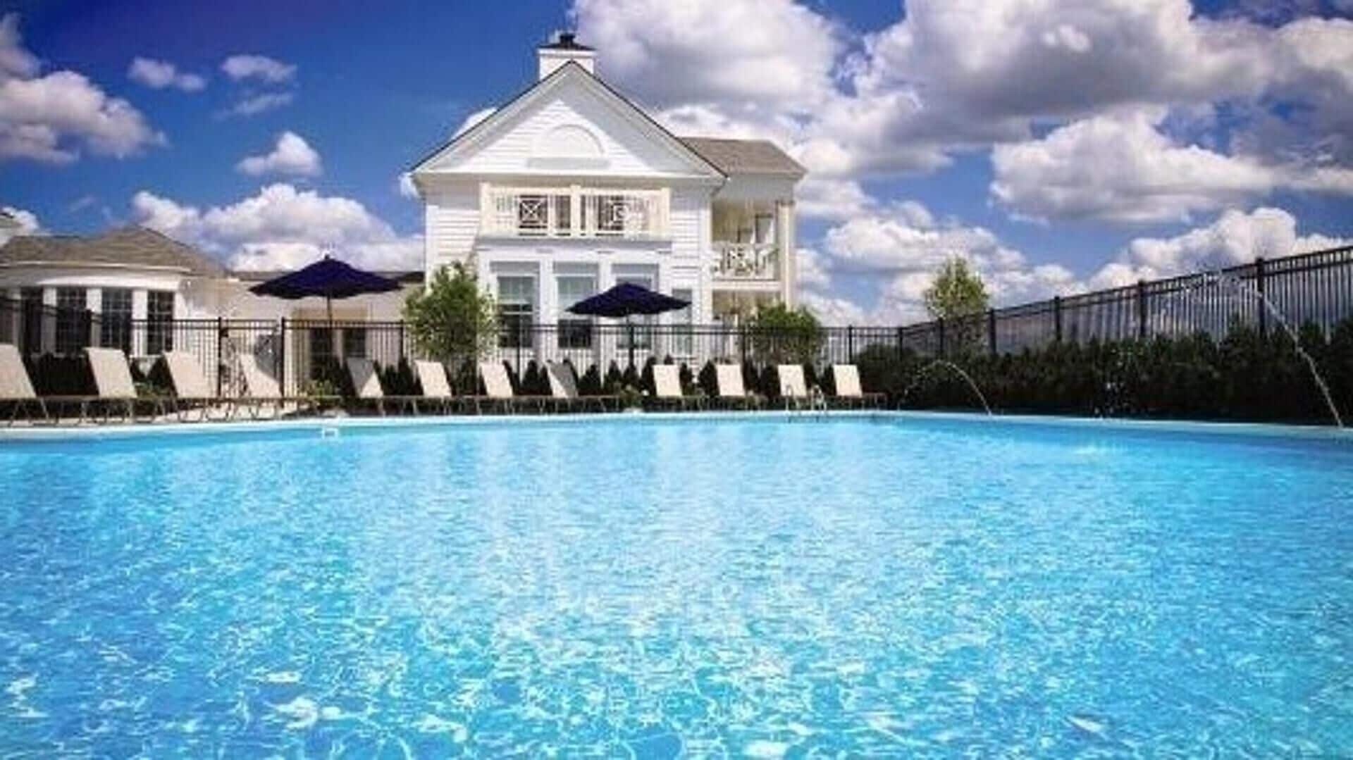 Ein Swimmingpool mit Liegestühlen und Sonnenschirmen, umgeben von einem Zaun und einem großen, weißen Gebäude mit Satteldach.