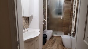 Et moderne badeværelse med hvid vask, toilet og bidet, brusebad med glasdør samt trægulv.