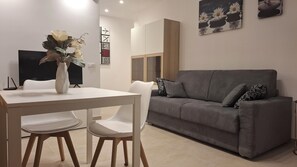 Ein modernes Wohnzimmer mit grauem Sofa, weißen Stühlen, einem weißen Tisch und einer Vase mit Blumen.