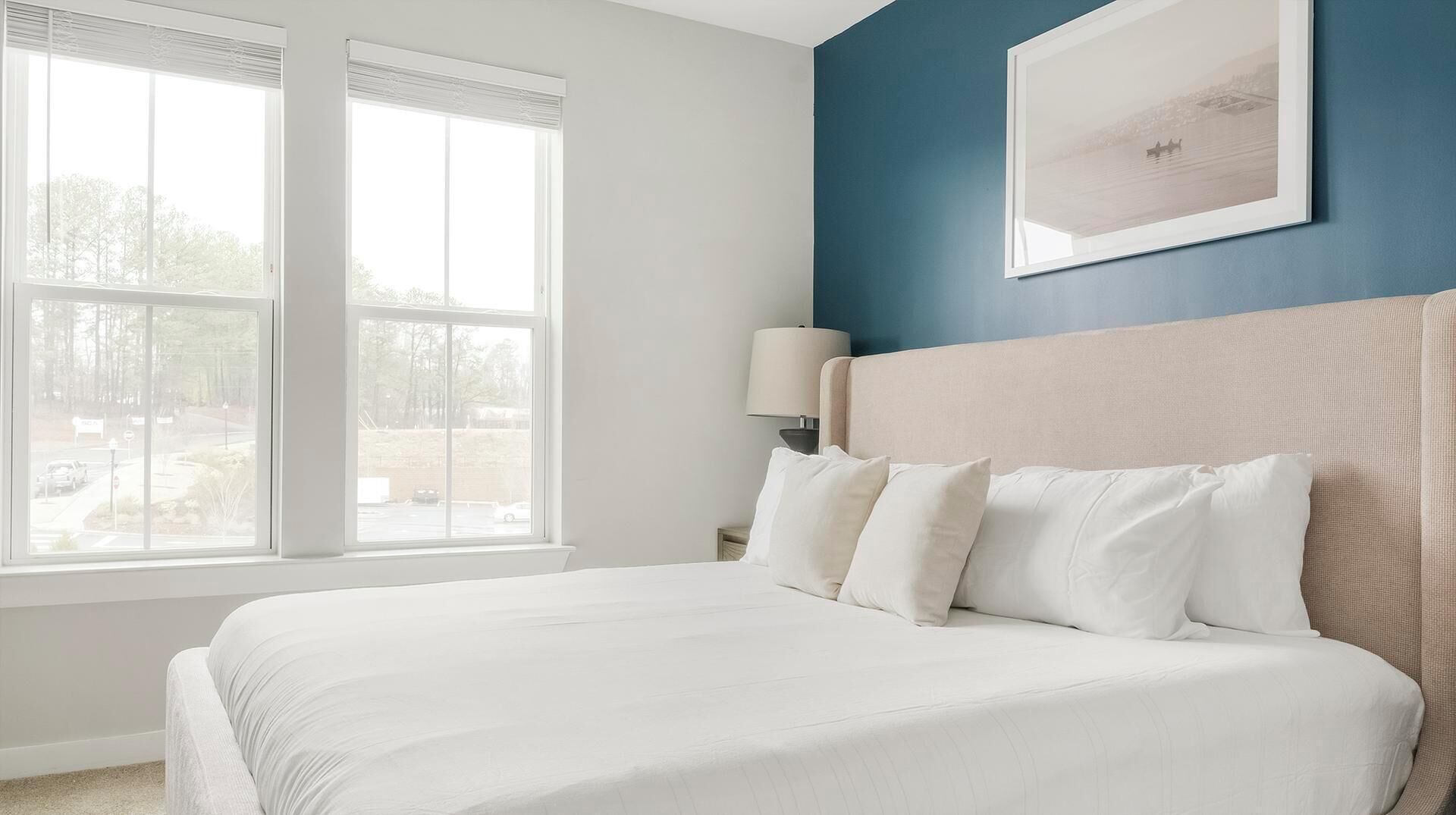 Une chambre d’hôtel avec un grand lit, deux fenêtres, une table de chevet, une lampe et un tableau encadré au mur.