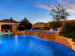 A swimming pool with a waterfall feature, surrounded by a landscaped garden and a building with a covered patio.