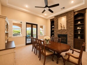 A dining room with a large table, chairs, a ceiling fan, and a stone fireplace.