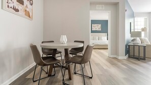 Een eetruimte met een ronde tafel en vier stoelen, een gang naar een slaapkamer, en een klein zithoekje met een lamp.