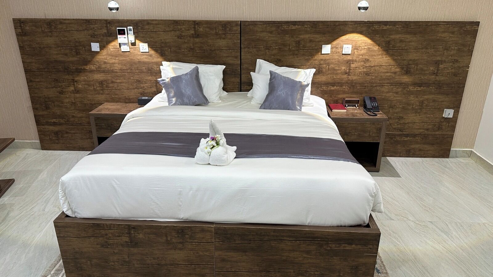 Een netjes opgemaakt bed met een houten hoofdeinde, een nachtkastje met een telefoon en een wandlamp.