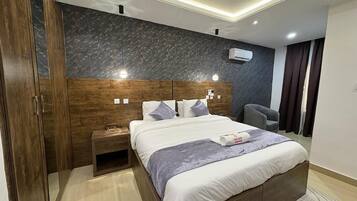 A modern hotel room with a large bed, wooden wardrobe, bedside table, and a built-in headboard.