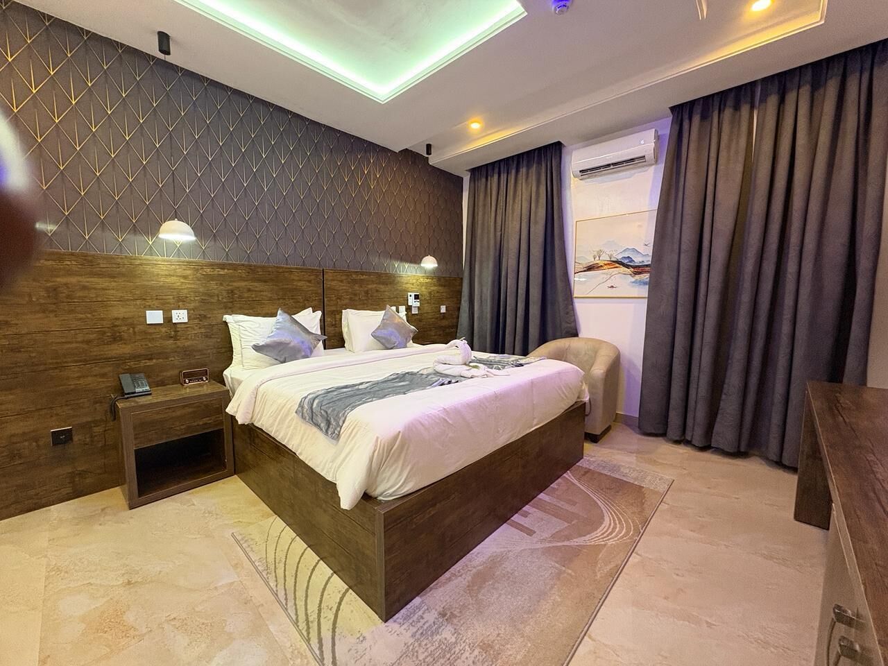 A modern bedroom with a wooden headboard, a bedside table with a lamp, a large bed with pillows, and a view of a mountain through the window.