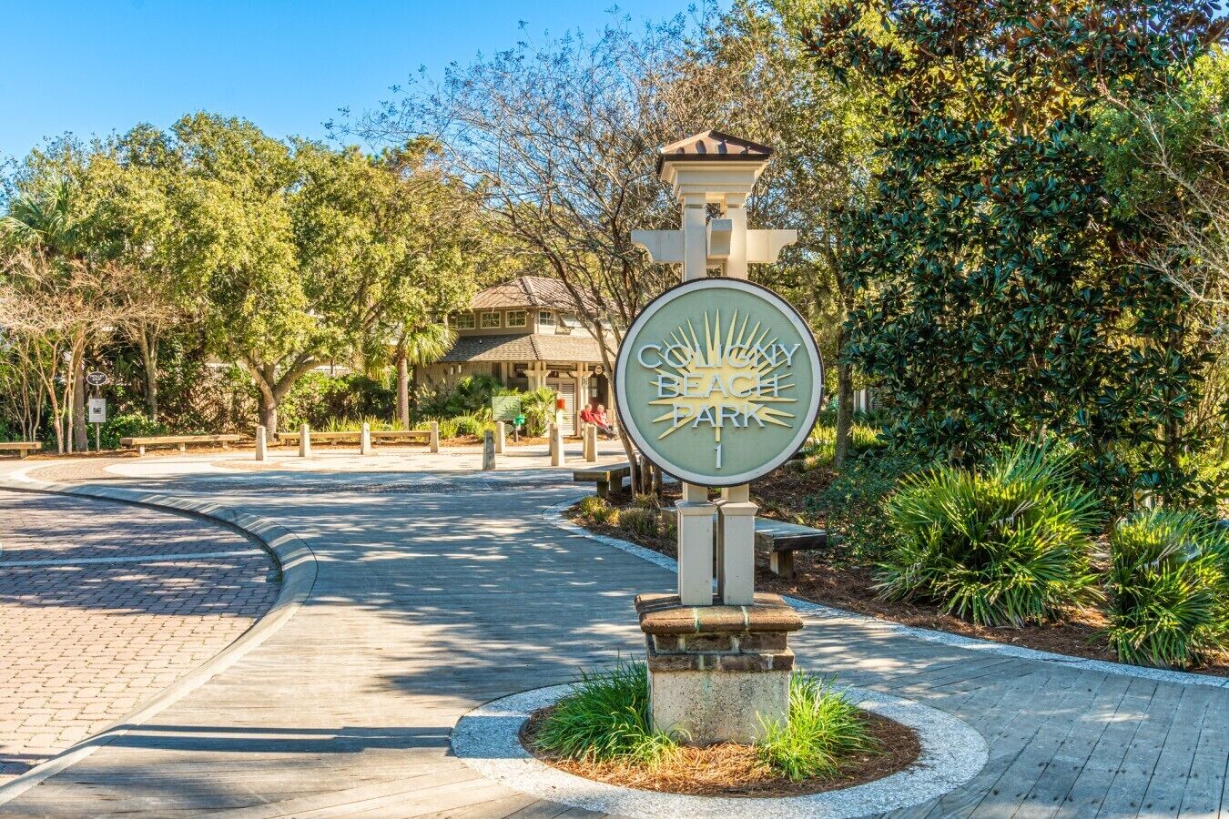 A park entrance sign with a sun motif, surrounded by greenery and a paved path.