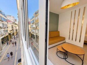 Ein modernes Interieur mit einer Couch und einem Tisch, Blick auf eine Straße mit Fußgängern und Häuser mit Balkonen.