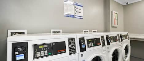 Una hilera de lavadoras industriales de carga frontal con pantallas digitales y paneles de control, en un lavadero limpio y bien iluminado con paredes grises y techo blanco.