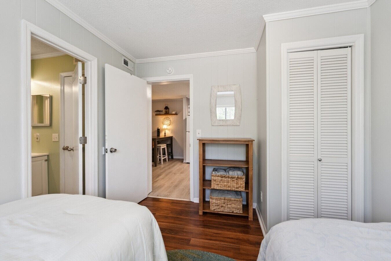 Une chambre avec deux lits, une étagère en bois avec des paniers, un petit bureau avec une chaise, et un couloir menant à une salle de bain.