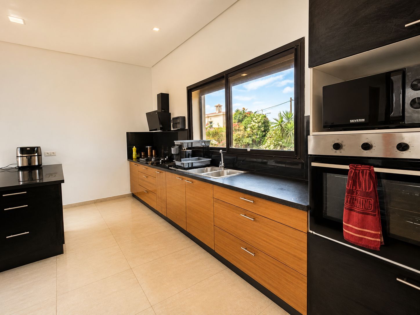 Una cocina moderna con encimera negra, armarios de madera, estufa de acero inoxidable y ventana con vistas al verdor.