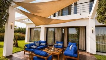 Un área de estar al aire libre y de estilo moderno con sofás y una mesa de centro, rodeada por un edificio blanco con ventanales grandes y una zona con sombra.