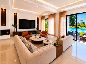 A modern living room with a flat-screen TV, comfortable seating, a fireplace, and a view of a swimming pool. - Palm Oasis Estate Dar Bouazza – 2 Private Villas • 16 Guests • 25m Swimming Pool (Tamaris)